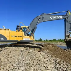 Żółta koparka Volvo EC240BLc pracuje na kamienistym brzegu rzeki. Zastanawiasz się, ile bierze koparka za godzinę?