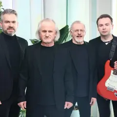 Czterech mężczyzn w czarnych strojach. Jeden z nich trzyma czerwoną gitarę.