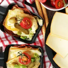 Deux poêlons de raclette fondue, garnie de tomates, cornichons et olives, accompagnés de fines tranches de fromage. Une variante gourmande !