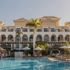 Piscina y edificio del Gran Meliá Palacio de Isora, con palmeras y tumbonas bajo el sol.