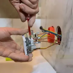 Elektryk montuje gniazdko, podłączając przewody zgodnie ze schematem. Widać fragment instalacji elektrycznej.