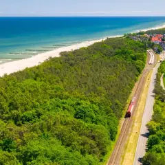 Czerwony pociąg jedzie wzdłuż wybrzeża. W tle widać plażę, morze i nadmorską miejscowość. Idealna podróż pociągiem nad morze.