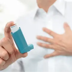 Mann hält Inhalator mit ipratropiumbromid. Seine andere Hand liegt auf der Brust, was auf Atemnot hindeutet.
