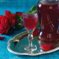 Aromatic nalewka z płatków róży w kieliszku i karafce, obok czerwona róża i płatki na srebrnej tacy.