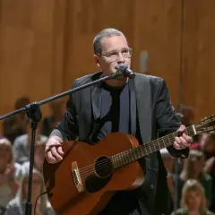 Artysta z gitarą i mikrofonem na scenie, przygotowuje się do koncertu w ramach **Męskie Granie 2026**. Publiczność w tle czeka na występ.
