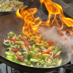 Ogień płonie nad warzywami w woku, tworząc aromatyczny stir fry. W tle widać smażone ziemniaki.