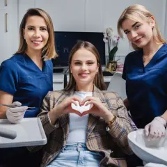 Uśmiechnięta pacjentka tworzy serce dłońmi, otoczona przez dwie dentystki w gabinecie stomatologicznym.