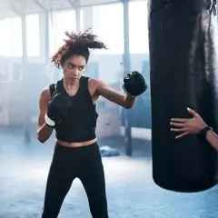 Woman training boxing for fitness at home, punching a heavy bag while a trainer holds it steady.