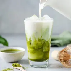 Eisiger Matcha Latte wird mit Milch übergossen. Die Zutaten für den Matcha Latte (kcal) sind auf dem Tisch verteilt.