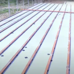 Drewniane legary ułożone na białej powierzchni, przygotowane do mocowania do betonu. Widać elementy mocujące.