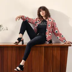 Mujer con rizos sentada en mueble de madera, luciendo distintos tipos de estilos de vestir: chaqueta floral, camiseta con brillos y jeans negros.