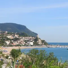 Vista panoramica della Riviera del Conero con un borgo arroccato su una collina, spiaggia attrezzata e un porto turistico.