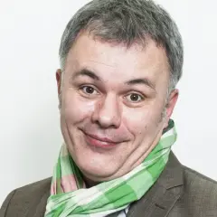Uśmiechnięty mężczyzna w kratkowanym szaliku, znany jako Mazurek z RMF FM, z szarymi włosami i brązową marynarką.