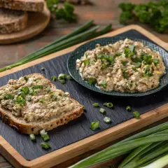 Pyszna pasta z tuńczyka na kromce ciemnego chleba, posypana szczypiorkiem. Obok talerz z pastą i świeże dymki.