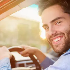 Un homme souriant au volant de sa voiture, prêt pour sa previsite contrôle technique. Le soleil brille, annonçant une journée sereine.