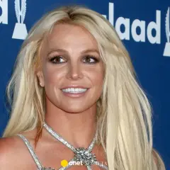 Uśmiechnięta Britney Spears, której wiek nie jest widoczny na zdjęciu, w błyszczącej kreacji na tle niebieskiej ścianki z logo GLAAD.