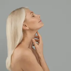 Äldre kvinna med blont hår rör vid sin hals, en bild som kan associeras med en kalkonhals operation.