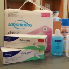 Pflegehilfsmittel Liste: Ein Paket Einlagen, zwei Schachteln Latex-Handschuhe, Desinfektionsmittel und ein Spray.