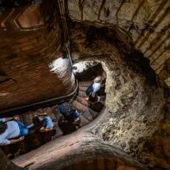 Persone esplorano le antiche cantine storiche di Montepulciano, scendendo una scala a chiocciola in mattoni e tufo.