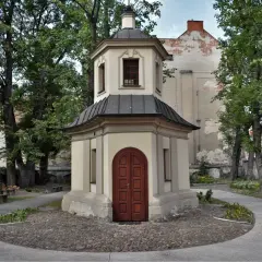 Kapliczka z drewnianymi drzwiami, otoczona drzewami i brukowanym placem. Jedna z urokliwych kaliskich atrakcji.