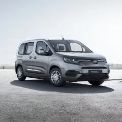 Srebrna toyota 7 osobowa czeka na pasażerów przy lotnisku. Ludzie z walizkami idą w stronę samochodu.