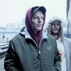 Dwóch mężczyzn w kapturach i czapkach, wyglądają jakby szli na jakieś seanse. Inni ludzie w tle.