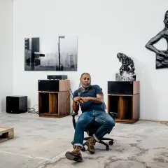 A man sits in an office chair in a gallery space, surrounded by art and audio equipment, a scene that feels like a snapshot of Arthur Jafa's creative process.