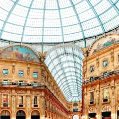 Inside Milan's Galleria Vittorio Emanuele II, a stunning glass dome arches over ornate buildings adorned with frescoes, reminiscent of grand Milan art galleries.