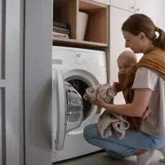 Une mère porte son bébé en écharpe tout en chargeant le linge dans le lave-linge. Une astuce de grand-mère pour éviter la mauvaise odeur du lave-linge.