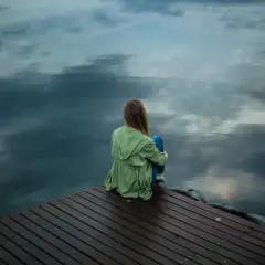 Ein Mädchen sitzt allein auf einem Holzsteg am Wasser. Sie wirkt nachdenklich, als ob sie nicht alleine sein kann.