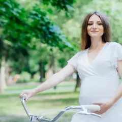Uśmiechnięta ciężarna kobieta w białej sukience trzyma kierownicę roweru w parku.