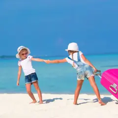 Due bambine si tengono per mano su una spiaggia tropicale, una tira una valigia rosa. Inizia la loro vacanza famiglia mare.