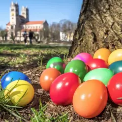 Bunte Ostereier liegen im Gras vor einem Baum, im Hintergrund eine Kirche. So feiert man an Ostern die Auferstehung.