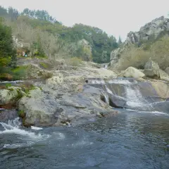 A fervenza de Segade, con auga caendo sobre rochas e un pequeno refuxio na ladeira verde.