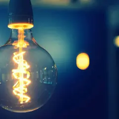 Żarówka z widocznym, rozgrzanym żarnikiem tworzącym spiralny strumień świetlny. W tle rozmyte, ciepłe punkty światła.
