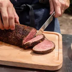 Un chef tranche un rôti de filet mignon parfaitement cuit sur une planche en bois.
