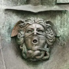 Brązowa rzeźba anioła z opaską na oczach i ustach, z wężami u dołu.