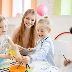 Uśmiechnięta przedszkolanka z dziećmi maluje. Ile zarabia przedszkolanka w prywatnym przedszkolu? Dzieci tworzą, a ona nadzoruje.