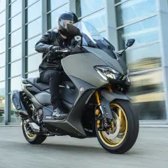 Hombre en moto clásica moderna, con chaqueta de cuero, circulando frente a un edificio de cristal.