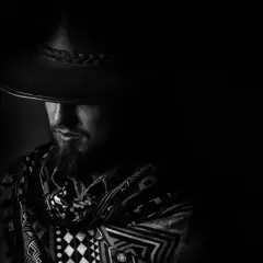 Mężczyzna w kapeluszu i wzorzystym szalu, z twarzą skrytą w cieniu. Dramatyczne chiaroscuro podkreśla tajemniczość postaci.