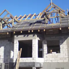 Budowa domu w toku. Drewniana konstrukcja dachu czeka na wiechę. W tle błękitne niebo.