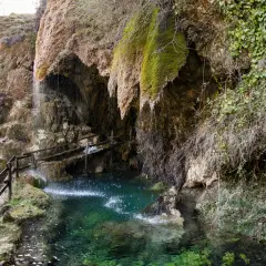 Cascata che scende tra rocce muschiose, formando una pozza d'acqua cristallina. Un sentiero con staccionata in legno conduce alle grotte di Labante.