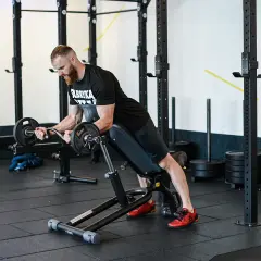 Mężczyzna z brodą wykonuje spider curl na ławce treningowej, podnosząc sztangę.