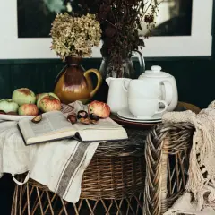 A cozy scene with apples, chestnuts, and an open book on a linen cloth, embodying the cottagecore aesthetic. A teapot and cups sit nearby, with a knitted blanket draped over a wicker chair.