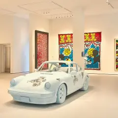 A white Porsche, artfully distressed, sits in a gallery. Vibrant Keith Haring-esque artworks adorn the walls, making for excellent Moco Museum reviews.