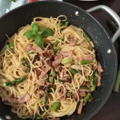 Pyszne spaghetti ze szparagami i boczkiem, udekorowane listkiem bazylii, czeka w patelni. Obok leżą świeże szparagi i czosnek.