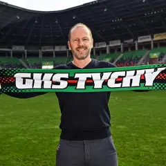 Uśmiechnięty mężczyzna, nowy trener GKS Tychy, trzyma szalik klubowy na tle stadionu.
