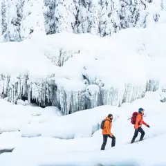 Två personer med ryggsäckar vandrar på vintern i ett snöigt landskap med istappar.