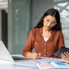 Kobieta w okularach pracuje przy biurku, licząc coś na kalkulatorze i notując. Widać laptopa i dokumenty. Wygląda na to, że ma sporo nadgodziny.