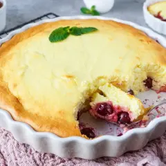 Ein goldbrauner Quarkauflauf mit Kirschen, garniert mit Minze, wird aus einer weißen Auflaufform serviert.